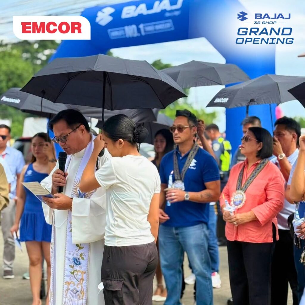 Priest reading a blessing under umbrellas as guests and staff gather outside the Bajaj 3S Shop