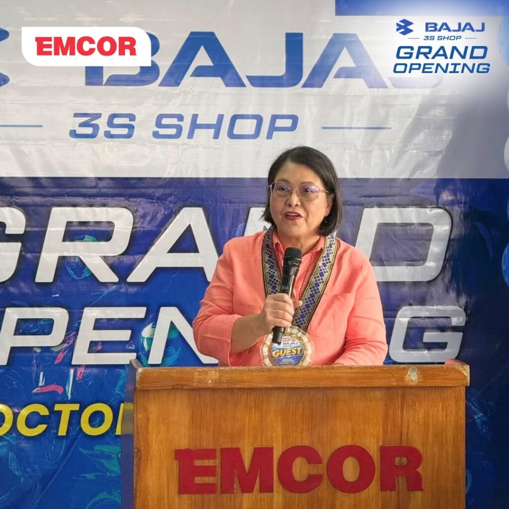 Guest in a coral blazer speaking at an EMCOR podium in front of a Bajaj 3S Shop Grand Opening backdrop